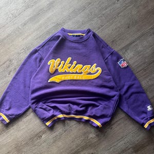 May include: A purple Vikings sweatshirt with yellow lettering and trim. The word "Vikings" is prominently displayed across the chest in a stylized font, with "FOOTBALL" underneath. An NFL logo patch is on the sleeve.