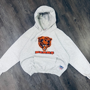 May include: A light gray hoodie featuring the Chicago Bears logo in orange and navy blue. The word "BEARS" is printed below the logo. The hoodie has a front pocket and a hood with a drawstring.