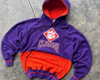 May include: A purple and orange Clemson University hoodie. The hoodie features a large front pocket, a drawstring hood, and the word "CLEMSON" in white script below a paw print logo.