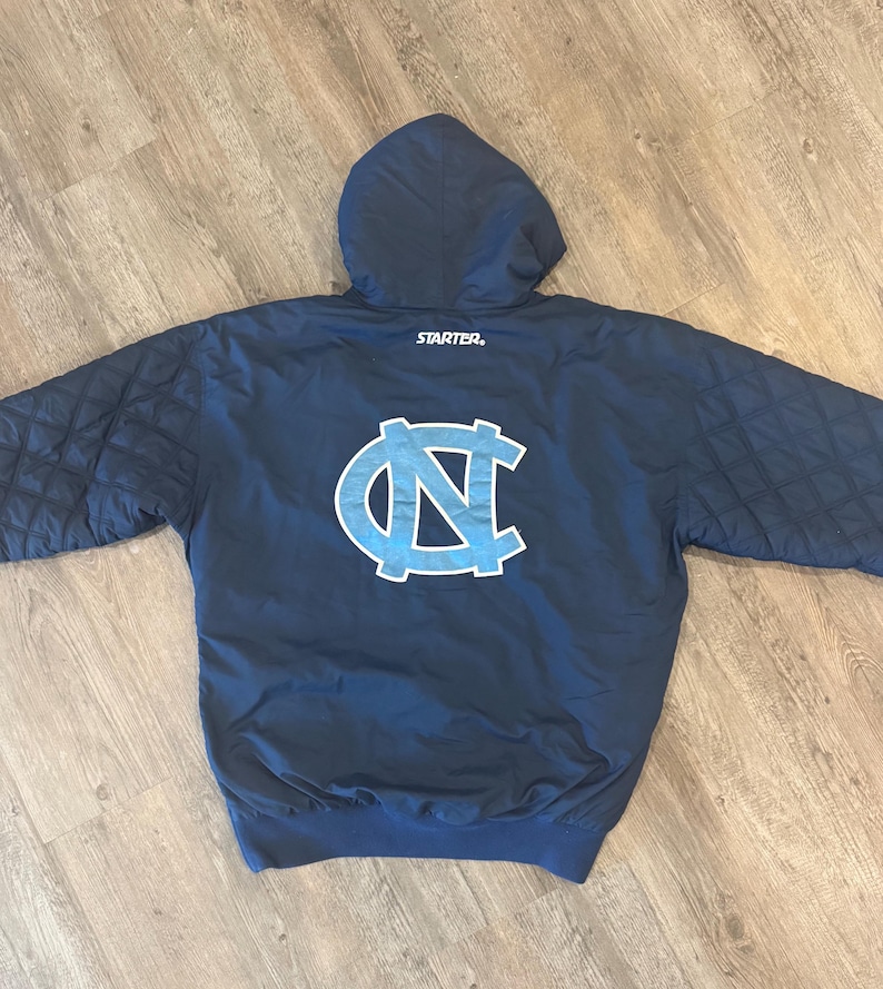 May include: Navy blue hooded jacket with a light blue and white logo on the back. The jacket has a quilted pattern on the sleeves and the Starter logo is visible. The jacket is made of a smooth, possibly nylon, material.