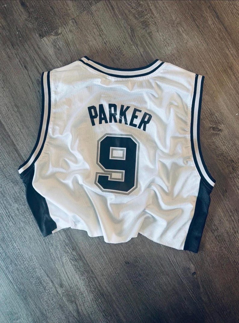 Puede incluir: Una camiseta de baloncesto sin mangas de malla blanca con ribetes negros en el cuello y las sisas. La parte posterior muestra el nombre PARKER en letras negras sobre el n&uacute;mero 9, impreso en negro con un contorno gris sobre la tela.
