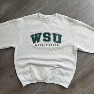 May include: A light gray heathered crewneck sweatshirt featuring the dark green text WSU and the words WRIGHT STATE printed in smaller dark green letters below. The garment has long sleeves and a Champion brand label at the inner neckline.