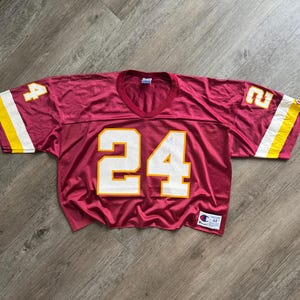 May include: A burgundy short-sleeved jersey with a V-neck collar. The front features the number 24 in large white numerals with yellow outlines. Each sleeve displays a white and yellow stripe and the number 4 on the right and 2 on the left.