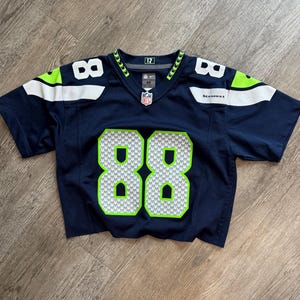 May include: A dark navy blue Seattle Seahawks jersey with short sleeves, featuring the number 88 in white and neon green on the front and shoulders. The jersey includes an NFL shield at the neckline, the text Seahawks, and a number 12 patch inside.
