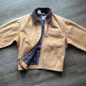 May include: A tan work jacket with a brown corduroy collar. The jacket has a zip closure, multiple pockets, and a blue and red striped interior lining. The Carhartt logo is visible on the tag.