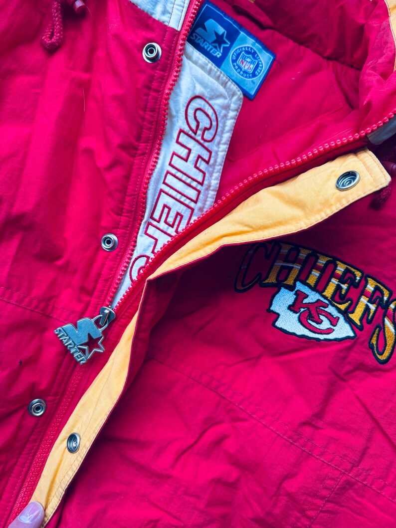 May include: Red and yellow Starter brand jacket with the Kansas City Chiefs logo and "Chiefs" written on the front. The jacket has a zipper and a snap closure.