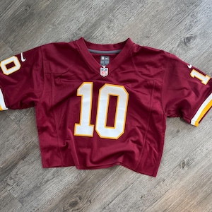 May include: A burgundy short-sleeved jersey featuring the number 10 in white with a gold outline on the front and sleeves. The shirt includes a Nike logo on the right shoulder, an NFL shield at the neckline, and a size L tag inside the collar.