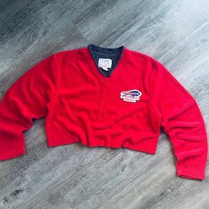 May include: A bright red long-sleeve V-neck sweater featuring a dark grey inner collar and a Buffalo Bills logo patch on the left chest. The garment has a cropped hemline and a white interior neck label with black text.