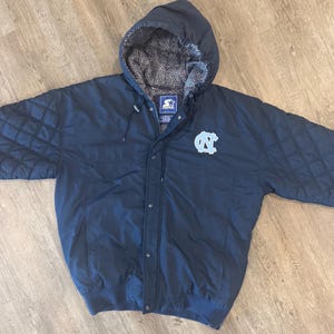 May include: Navy blue hooded jacket with a light blue logo on the chest. The jacket has a quilted design on the shoulders and a gray and white patterned lining in the hood. The jacket has a Starter logo.
