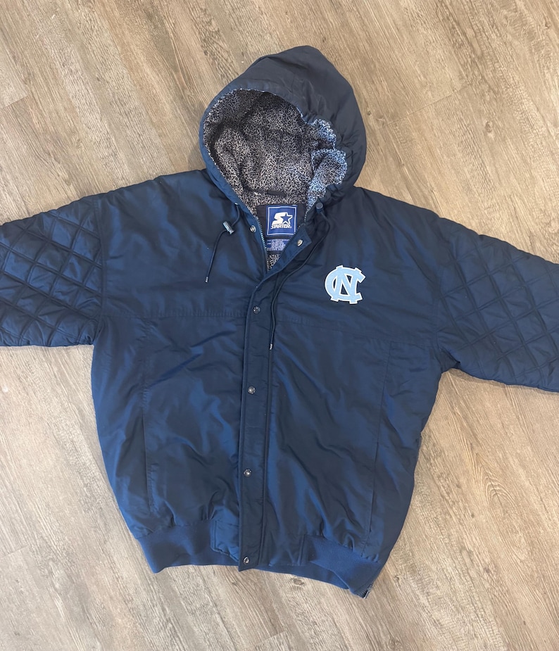 May include: Navy blue hooded jacket with a light blue logo on the chest. The jacket has a quilted design on the shoulders and a gray and white patterned lining in the hood. The jacket has a Starter logo.