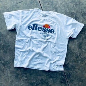 May include: White t-shirt with a blue "ellesse" logo and the text "The Best Looking Shoes in America".