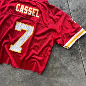 May include: A red football jersey with the name "CASSEL" in white letters with a yellow outline on the back. The number "7" is displayed in white with a yellow outline. The jersey has white and yellow stripes on the sleeves.