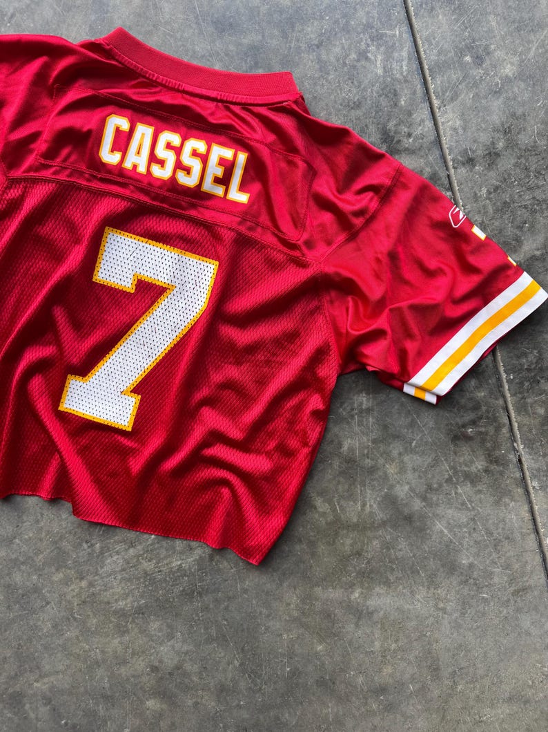 May include: A red football jersey with the name "CASSEL" in white letters with a yellow outline on the back. The number "7" is displayed in white with a yellow outline. The jersey has white and yellow stripes on the sleeves.