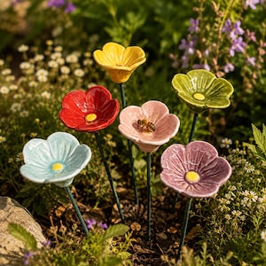 May include: A collection of six ceramic flower garden stakes in yellow, red, light blue, pink, green, and purple, each featuring a yellow centre and a thin green stem. A small bee is perched on the centre of the pink ceramic flower petal.