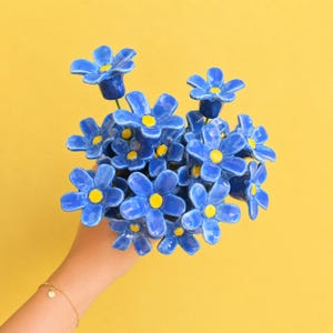 Puede incluir: Un ramo de flores de cerámica con pétalos azules de cinco puntas y centros de color amarillo brillante. Cada flor está unida a un tallo verde fino, creando un conjunto denso de piezas de cerámica azules y amarillas dispuestas juntas.