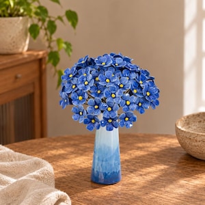 May include: A ceramic bouquet of small blue flowers with bright yellow centres arranged in a slender, tapered vase that features a gradient of light blue at the top transitioning to a deeper, saturated blue at the base, creating a cohesive display.