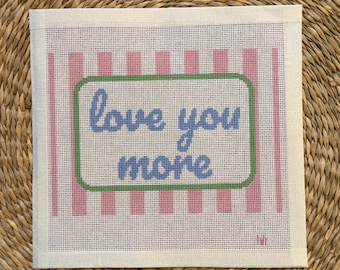 May include: A square needlepoint canvas featuring a pink and white vertical striped background. Centered is a rectangular frame with a green border containing the blue stitched text "love you more" in a cursive script font.