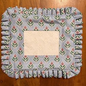May include: A rectangular light blue fabric placemat featuring a repeating floral pattern of pink flowers with green leaves and stems. The placemat has a ruffled border and a central rectangular white panel with a faint floral print.