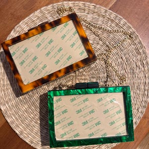 May include: Two rectangular acrylic box clutches with gold-tone chain straps. One clutch features a brown tortoiseshell pattern, while the other has a green marbled finish. Both have a central rectangular panel covered in 3M adhesive paper.