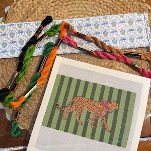 May include: A square needlepoint canvas featuring a leopard with a pink ribbon against a green and dark green striped background, accompanied by five skeins of embroidery floss in black, green, orange, pink, and brown, and a floral pouch.