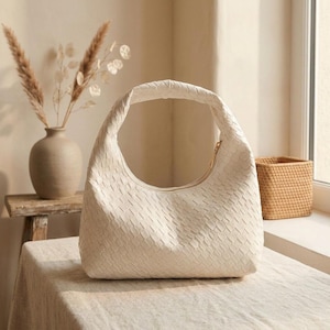 Könnte beinhalten: Eine cremefarbene Hobo-Tasche mit einer komplexen Flechtstruktur auf der gesamten Oberfläche. Die Tasche hat eine abgerundete, halbmondförmige Silhouette mit einem einzelnen integrierten Griff und einem sichtbaren Reißverschluss oben.