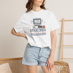 May include: A white short-sleeved t-shirt featuring a graphic of a vintage computer displaying a spreadsheet, a spiderweb, a ghost, and an orange pumpkin. The text reads SPOOKINESS & SPREADSHEETS in dark blue lettering centered on the chest.