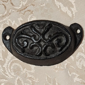 May include: A black cast iron drawer pull featuring an oval shape with a raised, symmetrical scrollwork pattern. The hardware includes two side mounting tabs, each with a single circular hole for attachment to a surface, providing a functional grip.
