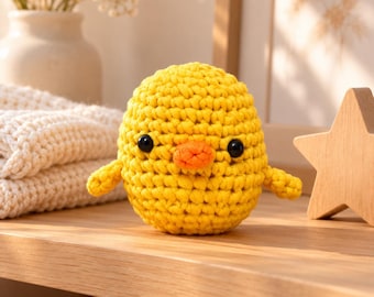 May include: A small, round, yellow crochet chick toy with a textured yarn surface, two black plastic bead eyes, and a small orange stitched beak. The handmade stuffed figure has a plump, spherical body and two tiny wings on its sides.