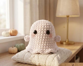 May include: A cream-coloured crochet ghost plushie with a textured yarn surface, two large black plastic safety eyes, and soft pink blush accents on the cheeks. The ghost has small rounded arms and sits atop a thick, cream-coloured knitted pillow.