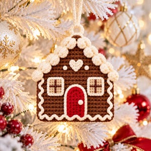 Puede incluir: Un adorno de casa de jengibre hecho de ganchillo en color marr&oacute;n con ribetes blancos estilo glaseado, una puerta roja y dos ventanas cuadradas. La casa presenta un peque&ntilde;o detalle de coraz&oacute;n blanco y una lazada a juego.