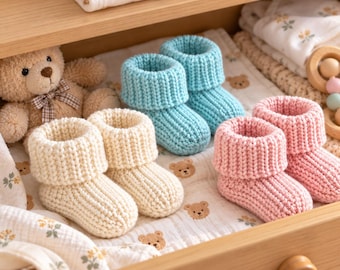 May include: Three pairs of knitted baby booties in cream, light blue, and soft pink, each featuring a ribbed texture and a folded cuff. The booties are arranged on a white fabric surface patterned with small brown teddy bear illustrations.