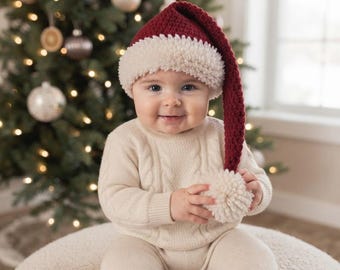 May include: A long, dark red crochet Santa hat with a thick, textured cream-coloured brim and a large cream pom-pom at the tip. The hat is worn with a cream-coloured cable-knit jumper and matching knit trousers, creating a festive holiday outfit.
