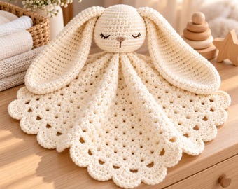 May include: A cream-coloured crochet security blanket featuring a plush rabbit head with long, floppy ears and closed eyes. The blanket body is circular with a scalloped edge and a textured, repeating shell stitch pattern throughout the design.