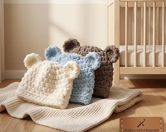 May include: Three chunky knit hats with bear ears in cream, light blue, and brown. The hats are made of thick, soft yarn and are arranged on a matching cream-coloured knit blanket. A brown leather tag displays the text dudecrochetexpert.