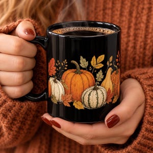 May include: A black ceramic mug featuring a colorful illustration of various pumpkins and autumn leaves in shades of orange, yellow, and cream. The design wraps around the exterior of the mug, which is filled with a dark liquid, likely coffee or tea.
