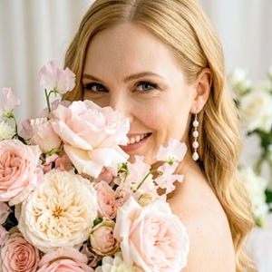 May include: A woman with blonde hair smiles, holding a bouquet of pink and peach roses and other flowers. She wears pearl drop earrings. The flowers are in various stages of bloom, creating a soft, romantic aesthetic.