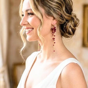 May include: A woman wearing long, dangling earrings with red beads. The earrings are attached to a gold chain and are clustered at the top, with individual beads cascading down. She is wearing a white dress.