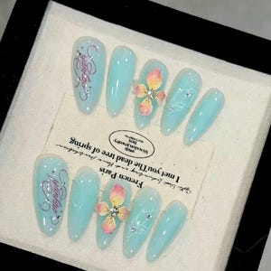Puede incluir: Un juego de diez uñas postizas en forma de almendra con acabado azul claro. El diseño incluye detalles florales rosas y amarillos, texto en plata, líneas blancas y pequeños brillantes transparentes. Presentadas sobre una tarjeta color crema.
