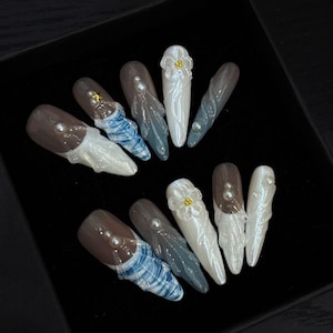 Puede incluir: Un juego de diez uñas postizas con una mezcla de diseños que incluyen detalles florales blancos en 3D, patrones de mármol azul y blanco, acabados en gris translúcido y pequeños adornos de perlas blancas con una estrella dorada en una uña.
