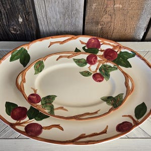 May include: Two oval ceramic serving platters with a cream-colored base, featuring a hand-painted design of red apples, green leaves, and brown branches. The rims of both platters are accented with a brown branch-like pattern and integrated handles.