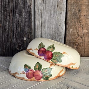 May include: Two cream-colored ceramic bowls stacked on top of each other. Each bowl features a hand-painted design of red plums on brown branches with green leaves encircling the exterior. The bowls have a smooth, glossy finish and rounded edges.