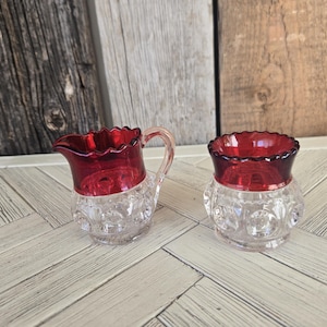 May include: A small glass creamer pitcher and a matching open sugar bowl featuring a clear base with a raised hobnail texture and a deep red stained glass rim. Both pieces have a scalloped edge along the top opening and a rounded body shape.
