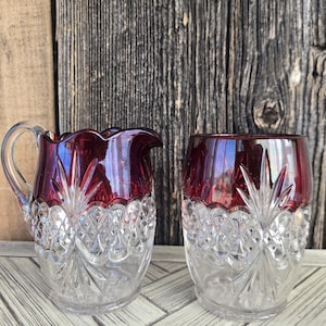 May include: A clear and cranberry red glass creamer pitcher and a matching tumbler. Both pieces feature a cut glass diamond pattern on the lower half and a smooth, deep red glass band around the top rim with vertical starburst cuts.