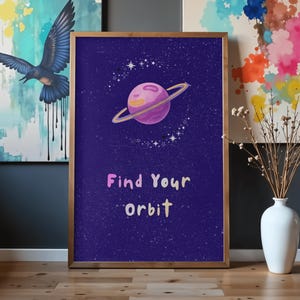 Könnte beinhalten: Ein gerahmtes, rechteckiges Wandbild mit einem dunkelblauen, sternenreichen Hintergrund, einem zentralen rosa Planeten mit Ring und kleinen weißen Sternen. Der Text "Find your orbit" ist in hellrosa und gelben Buchstaben darunter gedruckt.