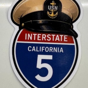 Puede incluir: Una pegatina que muestra una gorra de oficial naval USN de color beige y negro sobre una señal de tráfico de la Interestatal 5 de California en forma de escudo. La señal tiene una franja roja con INTERSTATE y una sección azul con CALIFORNIA 5.