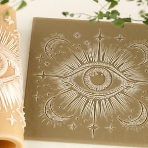 May include: A cylindrical tan rolling pin and a square tan flat tile, both featuring an embossed design of a central eye surrounded by radiating sunburst lines, stars, and crescent moons. The intricate pattern is carved into the surfaces.