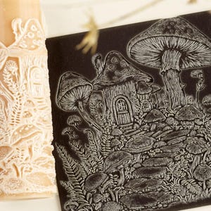 May include: A cylindrical beige rolling pin with a raised white relief pattern of mushrooms and foliage, paired with a square black tile featuring a white engraved illustration of a mushroom house, a stone path, and surrounding forest plants.