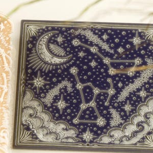 May include: A cylindrical beige rolling pin with a white embossed celestial pattern featuring stars and constellations, positioned next to a square dark blue tile displaying a matching white engraved design of a crescent moon, stars, and clouds.