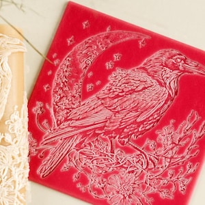 May include: A cylindrical beige patterned rolling pin and a square red silicone mat. Both items feature an embossed design of a raven perched on a flowering branch in front of a crescent moon surrounded by small stars, creating a detailed relief.