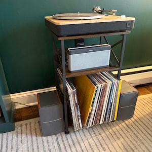 May include: A three-tier metal and wood record player stand featuring a turntable on the top shelf, a silver Bose speaker on the middle shelf, and a collection of vinyl records stored vertically on the bottom shelf with two speakers on the floor.